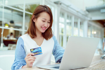 Portrait A beautiful Asian woman in a white shirt sitting in a bakery shopping online with her credit card and laptop computer in front of her hands.