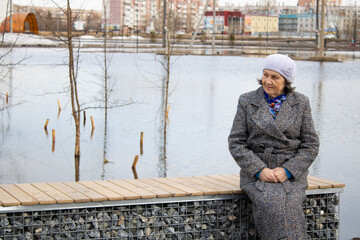 A lonely elderly woman is sitting on a bench. Floods in the city. The water spilled. Spring. The city was flooded with water. Grandmother next to the lake.