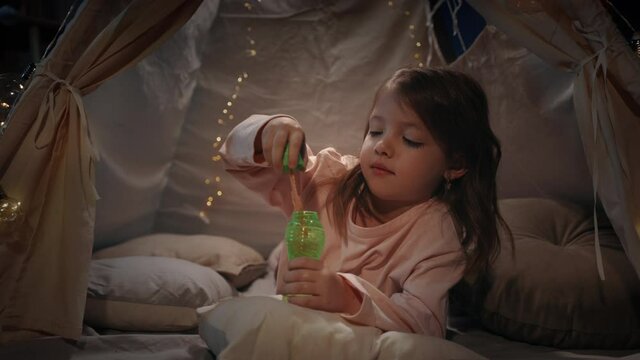 Adorable kid in cute makeshift blowing bubbless at home in evening. Beautiful little child lying on floor while spending good time. Concept of leisure and careless childhood.