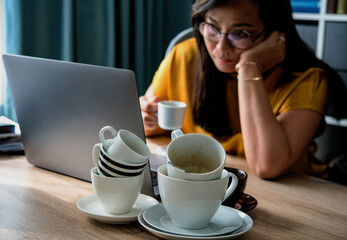 A piles of used coffee cups in front of an overtired Asian woman holding a cup of coffee sitting working frustrated exhausted looking at laptop screen. Caffeine addicted bad lifestyle concept. 