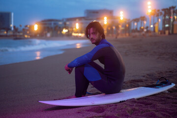 Night portrait of surfer in neoprene suit sitting on his surf board and looking at camera after surfing at Barcelona beach