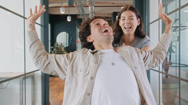 Young couple having fun as woman pushes man along walkway of modern open plan office in chair in slow motion