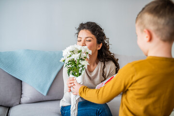 Selective focus of cute boy touching face of happy mom sitting in bed near flowers and mothers day card. Cheerful woman hugging boy and reading handmade greeting card with heart