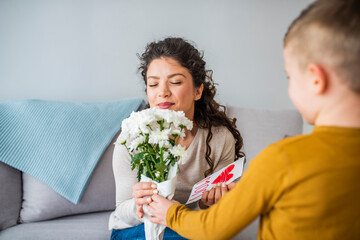 Selective focus of cute boy touching face of happy mom sitting in bed near flowers and mothers day card. Cheerful woman hugging boy and reading handmade greeting card with heart
