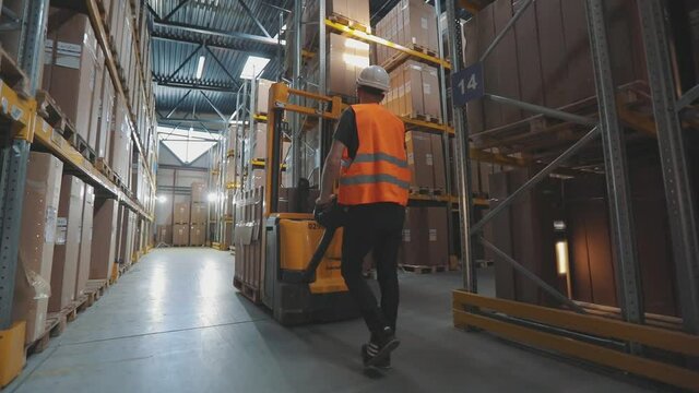 A worker walks through the warehouse with an electric hydraulic forklift. A worker transports cargo through the warehouse