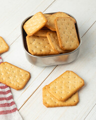 Cracker cookies in a stainless steel bowl with tablecloth on white wooden table background.