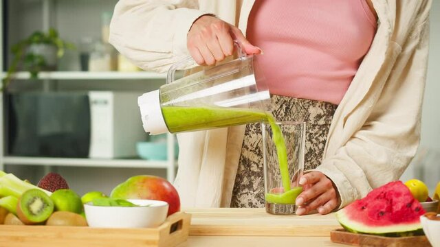 Young woman pouring green detox smoothie into glass from blender on table in the kitchen. Happy vegetarian cooking healthy food, fresh vegetable cocktail. Weight Loss Concept.