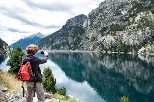 A hiker woman takes a photo with a mobile phone in a mountain lake, in the Cavallers reservoir, Boi Taull Valley, Lerida, Spain