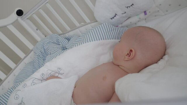 Baby boy sleeping in a crib with installed IP wireless security camera as baby monitor, interior of a modern apartment bedroom. High quality 4k footage