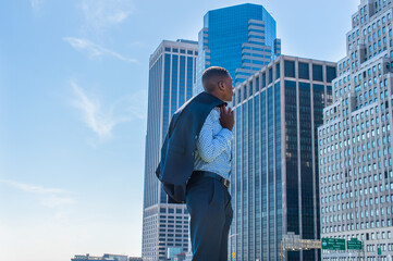 Taking off his jacket and putting on his shoulder, a young black businessman is standing in the front of a business district, hopefully looking at high buildings..