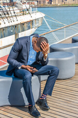 Dressing in a blue suit, leather shoes, a hand touching the top of his head , a young black businessman is sitting on the deck, looking down at his mobile phone.