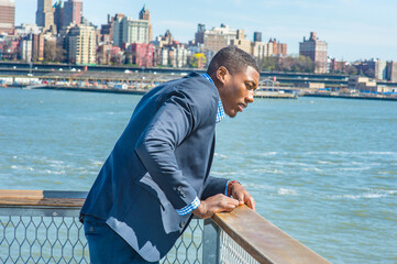 a young black businessman is standing by the river in New York City, looking down