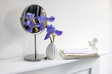 A branch of blue iris in a white fluted vase next to a round mirror, a figurine of an angel and a pile of white papers on a table against a white wall