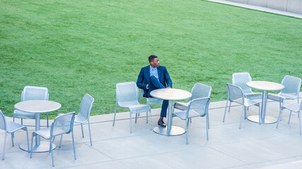 A young handsome black businessman is sitting on a chair by a green lawn and waiting for you..