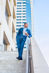 Holding a tablet computer, leaning on a railing, a young handsome black businessman is standing outside of a business building, reading, thinking and working.