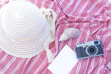Summer vacation still life. White straw hat, shell and camera on red and white towel. Blank invitation card or greeting card mockup. Concept of vacation and relaxation. Flat lay, top view.