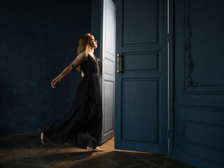 A young woman in a black dress opens a door from which light is pouring.