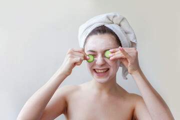 Girl with white facial mask holding cucumber slices isolated on white background. Woman applying white nourishing mask or creme on face. Skin care treatment spa natural beauty and cosmetology concept.