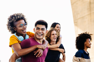 Diverse group of young people having fun together outdoors - Happy multiracial men giving girlfriends piggyback ride in city street - Friendship lifestyle concept - Focus on asian guy and afro woman