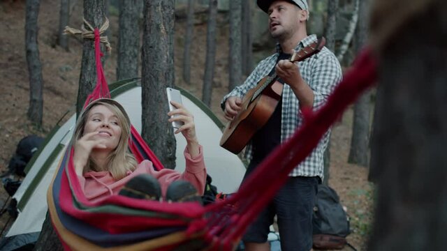 Young blonde is taking selfie with smartphone camera swinging in hammock in forest while boyfriend attractive guy is playing guitar having fun outdoors