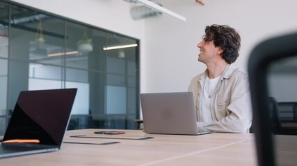 Casually dressed young man working on laptop at desk in modern open plan office is joined by female colleague in slow motion