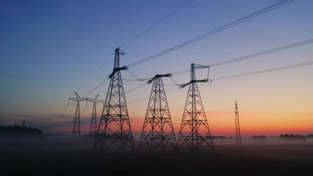 Aerial view high voltage steel power pylons in field covered with fog countryside. Misty early morning, dawn. Drone flight low over power transmission lines. Electric tower line, sunrise sunset