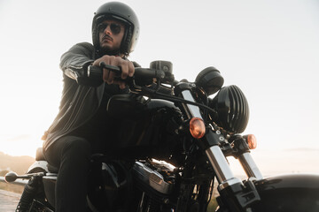 Fototapeta na wymiar Portrait of handsome biker sitting on his vintage motor bike, wearing leather jacket, helmet and sunglasses