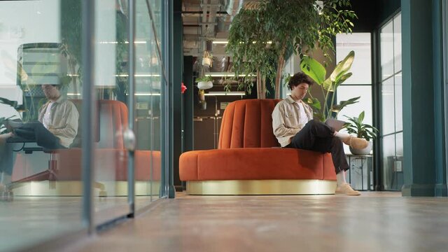 Full length shot of casually dressed young man working on laptop in modern open plan office sitting on orange circular sofa taking call on mobile phone