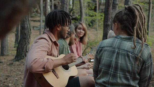 Slow motion of cheerful friends multi-ethnic group playing guitar and singing songs having fun outdoors in forest. Nature and vacations concept.