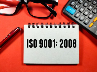 Fototapeta premium Quality assurance concept.Text ISO 9001:2008 writing on notebook with pen,glasses and calculator on red background