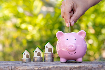 hand holding coin in pig piggy bank and house design on the pile of coins money concept Real estate...