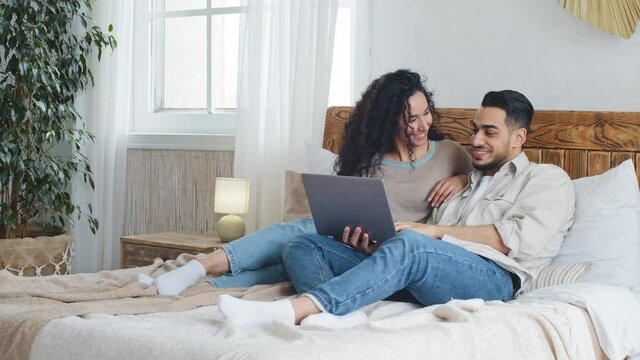 Spanish married couple arab ethnic handsome bearded boyfriend and curly girlfriend husband and wife lie together on bed in cozy bedroom use laptop looking at computer screen smile laughing talking