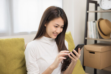 Happy young asian woman looking at screen of smartphone sitting on sofa in living room she shopping online. She very happy. Teenage girl reading news from mobile phone feeling excited.