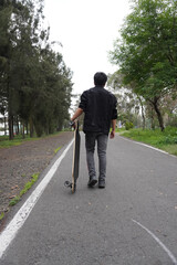 Young man with a longboard in a park. He is walking on the asphalt.