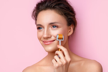 Beautiful woman using derma roller for her facial skin. Photo of woman on pink background. Beauty and skin care concept - obrazy, fototapety, plakaty