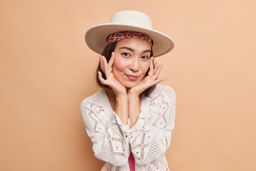 Natural beauty people and fashionn concept. Attractive Asian woman touches face gently looks at camera has pleased expression healthy skin wears hat and jumper isolated over beige background