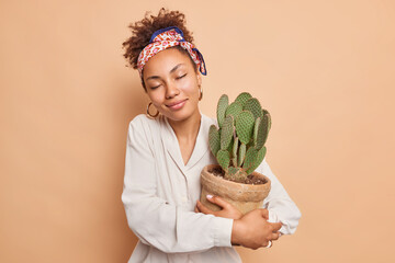 Indoor shot of happy dark skinned woman gets potted cactus as present fond of houseplants takes...
