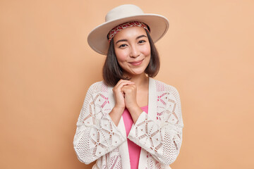 Portrait of lovely brunette Asian woman keeps hands together being touched by heartwarming words wears stylish hat white knitted jumper poses against beige background. Sincere human feelings