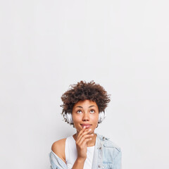 Fototapeta na wymiar Lovely young Afro American woman with thoughtful expression focused above has dreamy look listens lyrics songs via headphones concentrated upwards isolated over white background blank copy space