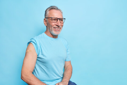 I got Covid 19 vaccine. Smiling bearded elderly man shows shoulder with band aid after vaccinating has happy expression wears t shirt spectacles isolated over blue background copy space for text