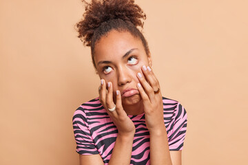 Dark skinned woman feels bored keeps palms on face has indifferent expression doesnt want to do wears casual striped t shirt isolated over beige studio wall being impatient cannot stand dull talk