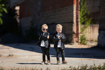 Blond twin boys in dark glasses and leather jackets.The little twins are stylishly dressed in a rock style.Cute kids look like celebrities.A children's photo on the background of an abandoned building - obrazy, fototapety, plakaty
