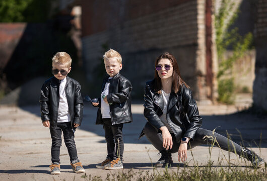 Mom and the twins in leather jackets and sunglasses.A bold and cool family image.Blonde little twin boys.The family is dressed in rock style.Mother and sons on the background of an abandoned building