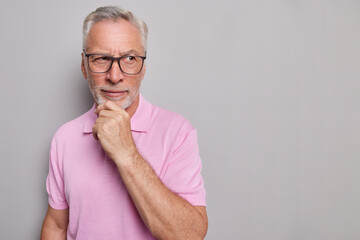 Horizontal shot of bearded thoughtful man holds chin looks pensively away being deep in thoughts...