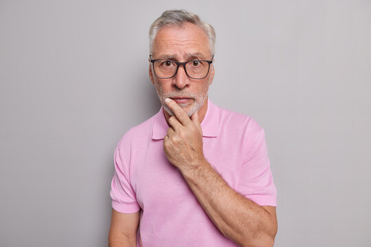 Serious bearded grey haired man looks attentively at camera holds chin has worried expression wears spectacles casual pink t shirt isolated over grey background. Human face expressions concept