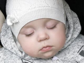 Portrait of a sleeping little baby wearing a creamy cap. Soft alabaster skin. Delicate baby dressed  for a walk. Calm healthy sleep outside. 