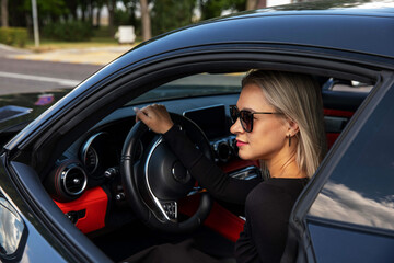 A blonde girl in sunglasses sits in the car and put her hand on the steering wheel