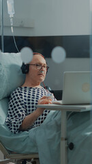 Hospital ward patient listening to music in intensive care room at clinic. Elder man using laptop and headphones to relax while receiving healing treatment to cure disease, illness