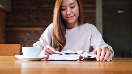 Obraz premium Closeup image of a beautiful asian woman holding and reading a book with coffee cup on the table
