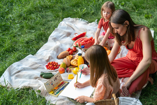 Mom spends time in nature with her lovely daughters. Drawing in park, picnic on blanket.